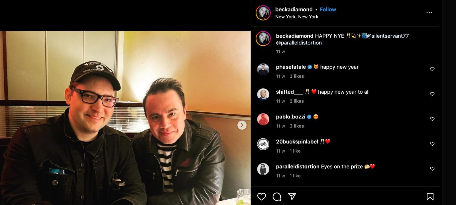 Screenshot of an instagram post from user @beckadiamond. At left, Dominick Fernow is wearing black plastic glasses, a black hat that says "Vatican Shadow" on it, a black jean hacket. Silent Servant has short dark hair visible, and is wearing a black leather jacket and a striped black-and-white shirt. They are seated close together, looking into the camera, and in the background are walls and a lamp casting light on the walls behind them