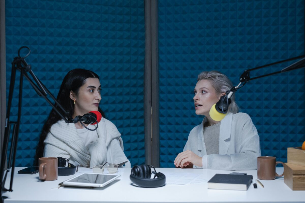 Two people sit in a podcast setting with mics. One is listening and the other is speaking with intensity.