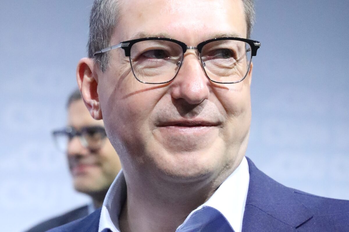 Alexander Dobrindt wearing glasses and a blue suit, standing in front of a blurred background with the logos of CDU and SPD partially visible.