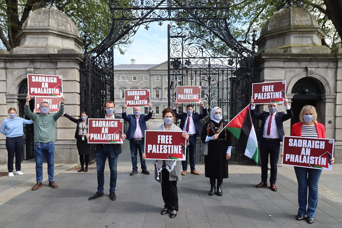 A group of people stand in front of a gate holding red signs saying "Free Palestine" in English and Irish. All are wearing masks with a keffiyeh pattern.