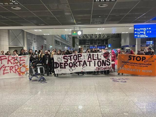 A crowd of protestors are holding up a anti deportation signs including a large banner that says "Stop All Deportations"